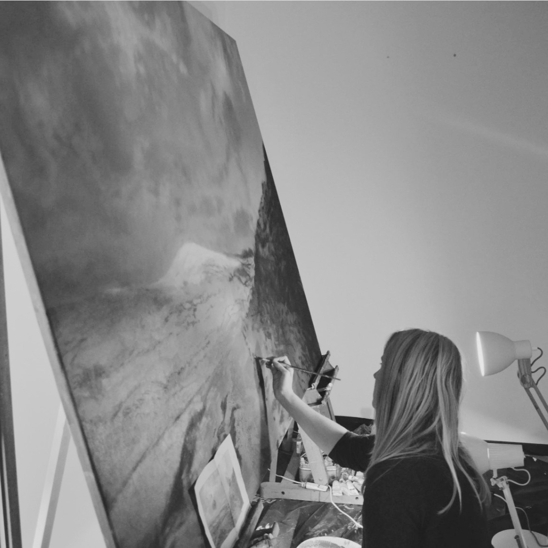 Black and white photograph of Southern Highlands artist Carla Gates painting a large-scale landscape artwork in her studio. Carla stands at an easel, carefully working on the dramatic, atmospheric piece using fine brushes. The studio setting is softly lit, with paints and materials surrounding her as she focuses on capturing light, texture and mood within the landscape.